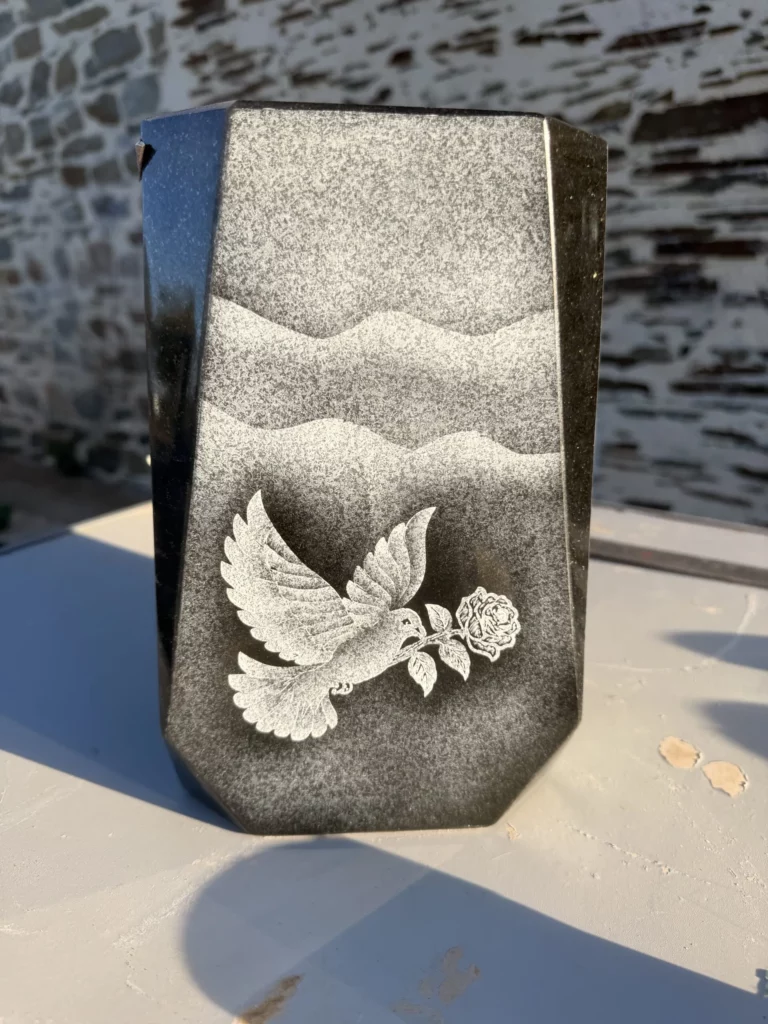 Colombe tenant une rose, gravée en grisé sur un vase en granit sous un paysage de collines.