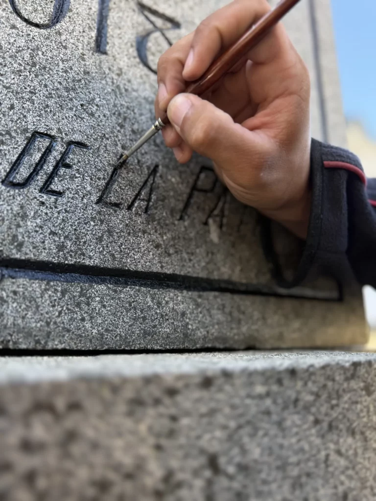 Main reprenant au pinceau fin des lettres gravées dans la pierre d’un monument.