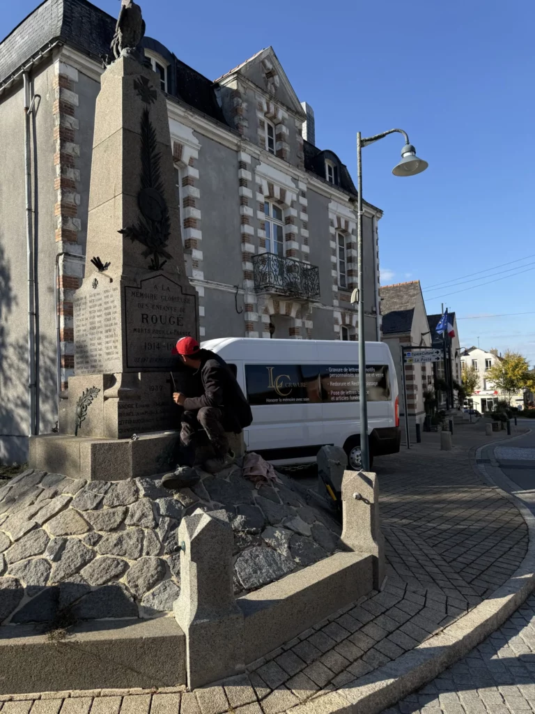 L’artisan au travail au pied d’un monument aux morts, sur une place de bourg.
